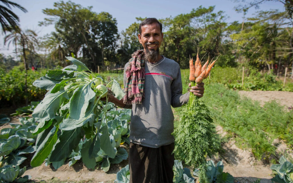 Een glimlachende man staat in een moestuin en houdt een grote bos vers geoogste wortelen en koolrabi omhoog.