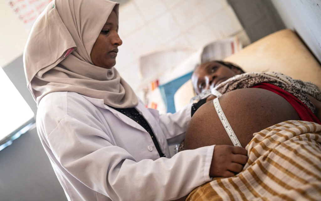 Een zorgverlener met een hoofddoek en een witte doktersjas meet met een meetlint de buik van een zwangere vrouw die op een onderzoekstafel ligt.