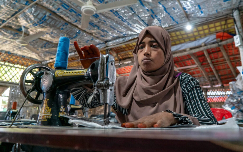 Een vrouw met een bruine hoofddoek werkt geconcentreerd achter een traditionele naaimachine in een lichte werkplaats.