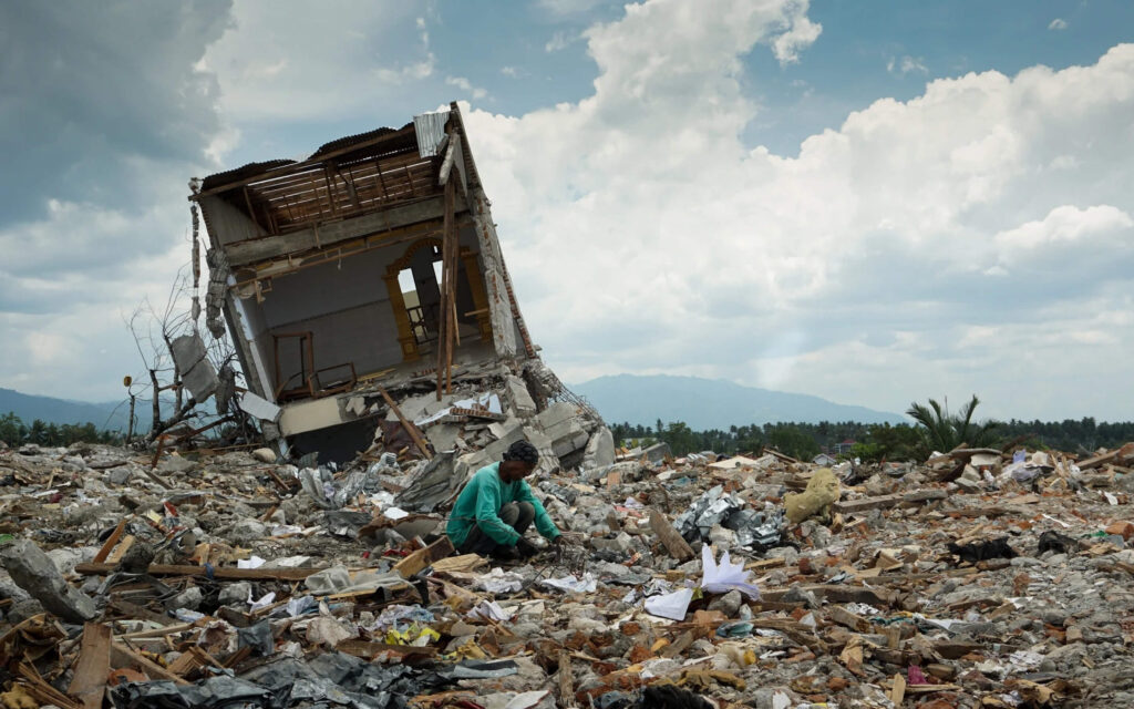 Een persoon zit gehurkt tussen de brokstukken van een volledig verwoest gebied, met op de achtergrond de restanten van een scheefgezakt gebouw na een natuurramp.