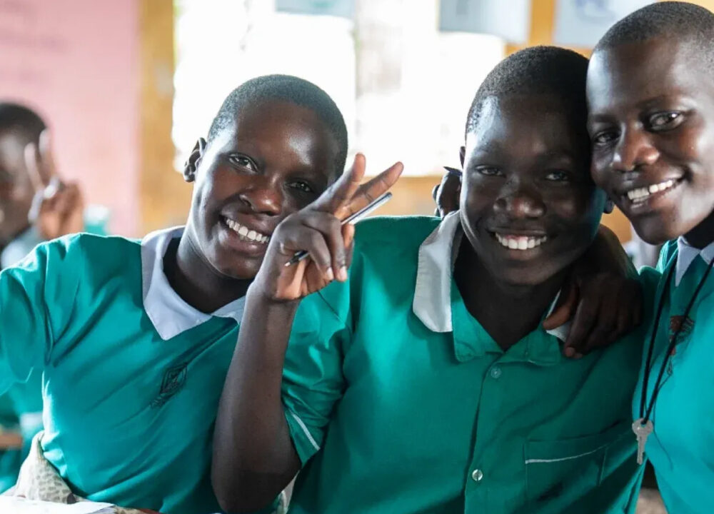 Drie lachende jongeren in groene schooluniformen poseren samen in een klaslokaal, waarbij één een vredesteken maakt.