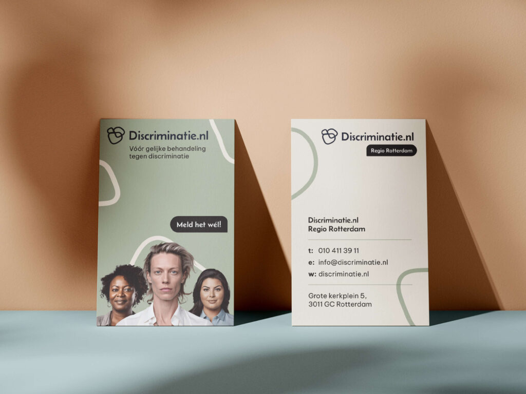 Twee visitekaartjes van Discriminatie.nl Regio Rotterdam tegen een zachtgekleurde achtergrond. Het ene kaartje is de voorkant met de slogan "Meld het wél!", het andere de achterkant met contactgegevens.