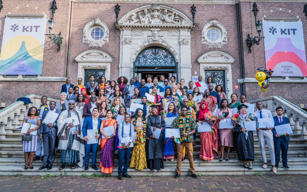 Een grote, diverse groep afgestudeerden in formele kleding en traditionele kledij poseert trots met hun diploma's op de monumentale trappen voor het KIT-gebouw in Amsterdam.
