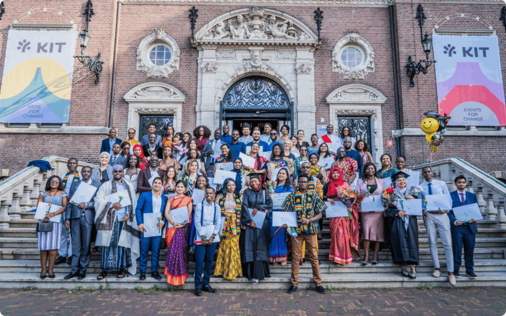 Een grote, diverse groep afgestudeerden in formele kleding en traditionele kledij poseert trots met hun diploma's op de monumentale trappen voor het KIT-gebouw in Amsterdam.