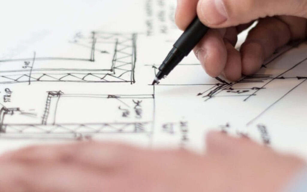 Close-up van een hand die met een zwarte fineliner nauwkeurige lijnen tekent op een technische bouwtekening of plattegrond.