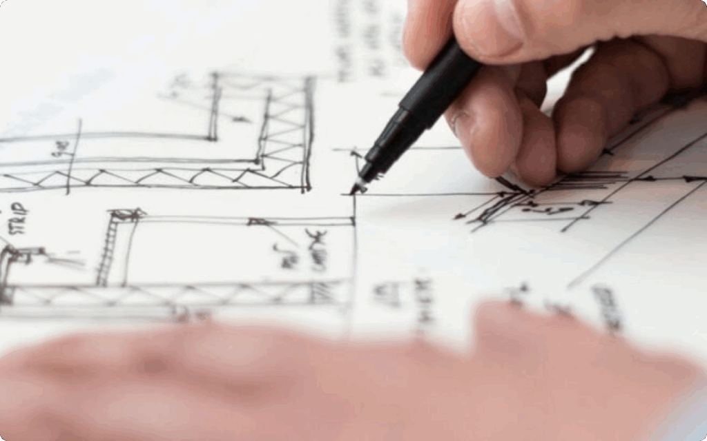 Close-up van een hand die met een zwarte fineliner nauwkeurige lijnen tekent op een technische bouwtekening of plattegrond.