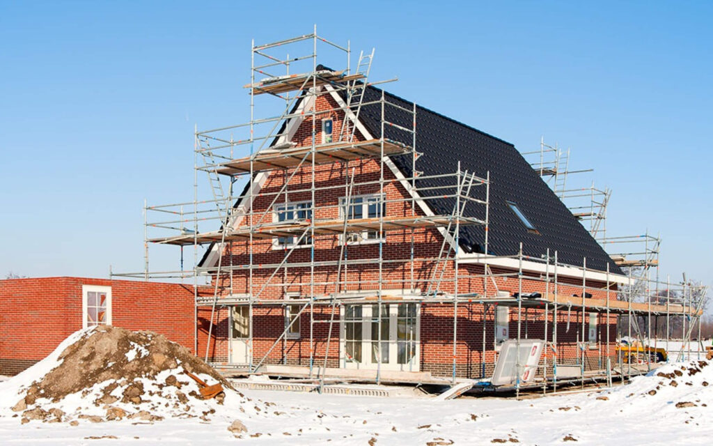 Een vrijstaand bakstenen huis in aanbouw, volledig omringd door steigers, op een bouwplaats bedekt met een dun laagje sneeuw onder een blauwe lucht.