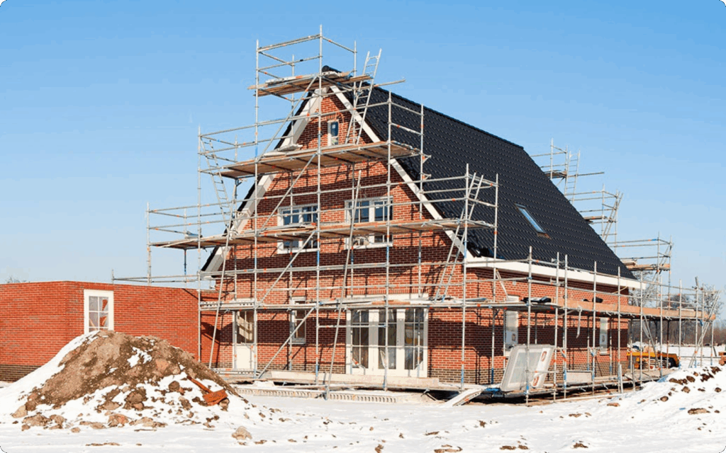 Een vrijstaand bakstenen huis in aanbouw, volledig omringd door steigers, op een bouwplaats bedekt met een dun laagje sneeuw onder een blauwe lucht.