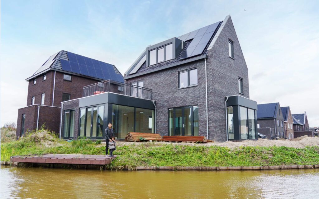 Een man en een klein kind staan op een vlonder bij de tuin van hun nieuw opgeleverde, moderne grijze bakstenen woning met zonnepanelen aan het water.