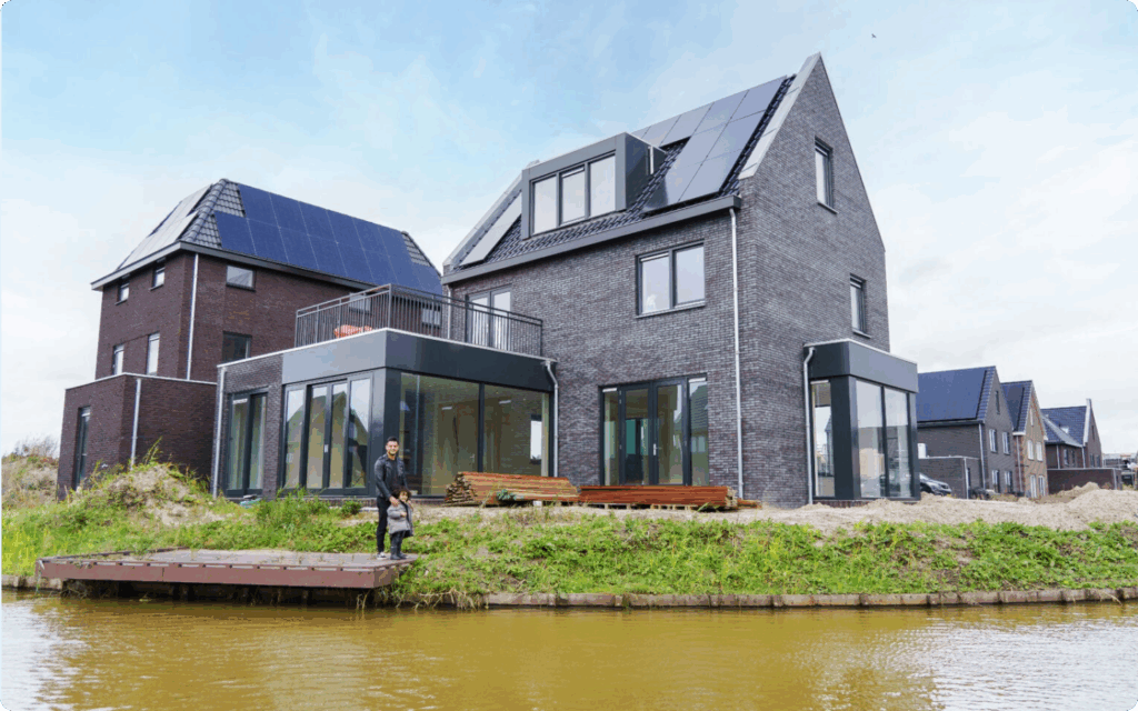 Een man en een klein kind staan op een vlonder bij de tuin van hun nieuw opgeleverde, moderne grijze bakstenen woning met zonnepanelen aan het water.