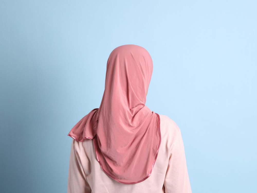 Achteraanzicht van een persoon met een roze hoofddoek en een bijpassend shirt tegen een lichtblauwe achtergrond.