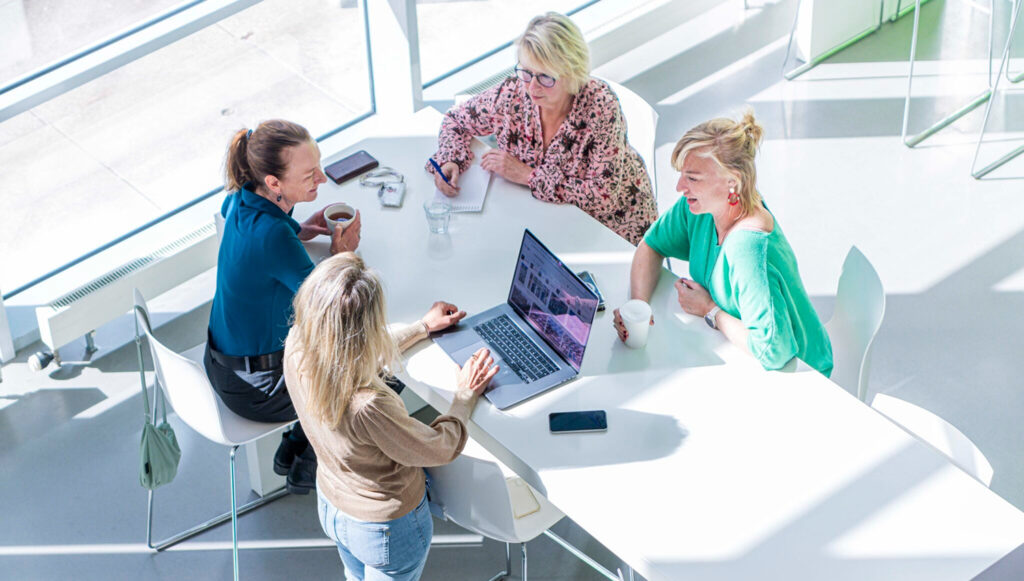 Bovenaanzicht van vier vrouwen die in overleg zijn aan een grote witte tafel in een licht, modern kantoor; een laptop staat open op tafel.