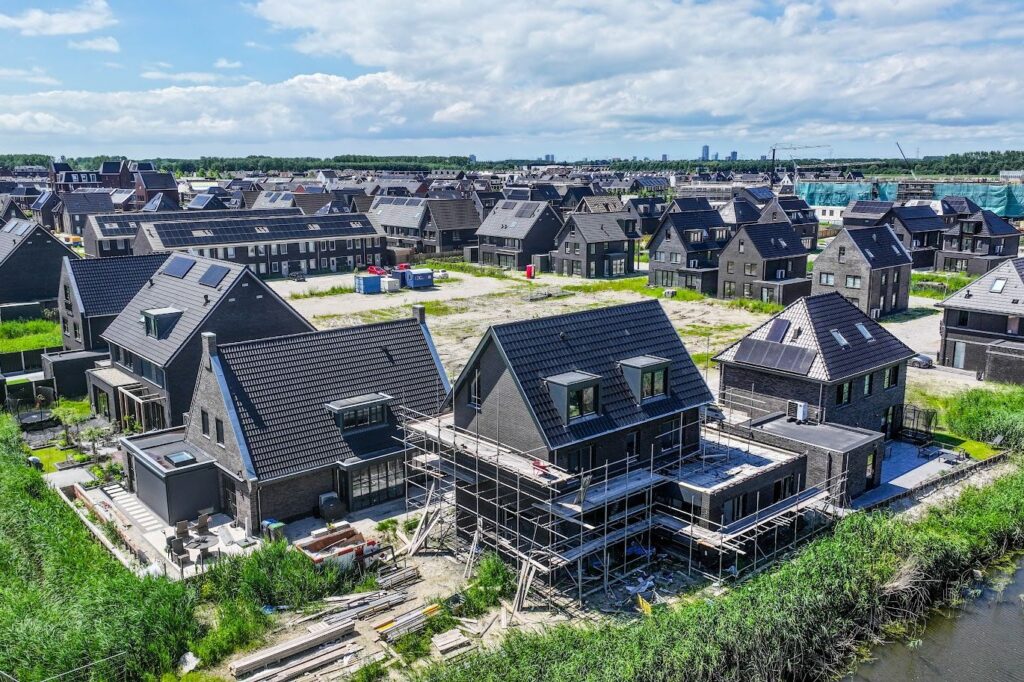 De afbeelding toont een luchtfoto van een nieuwbouwwijk in aanbouw, met een mix van voltooide en onvoltooide woningen en bouwmaterialen.