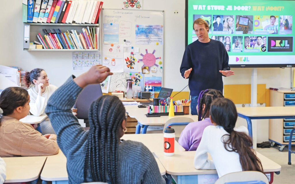 Een leraar staat voor een digibord in een klaslokaal en spreekt tegen een groep leerlingen die aan hun tafels zitten; een leerling steekt een hand op om een vraag te stellen.