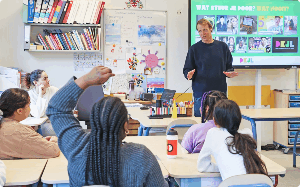 Een leraar staat voor een digibord in een klaslokaal en spreekt tegen een groep leerlingen die aan hun tafels zitten; een leerling steekt een hand op om een vraag te stellen.