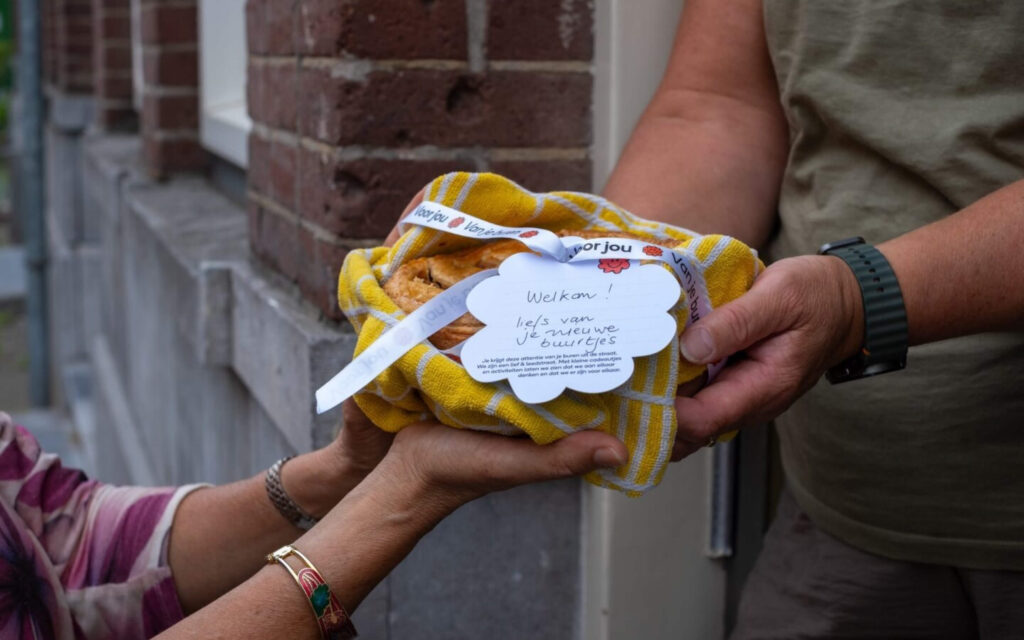 Een buur die een andere buur welkom heet in de straat, je ziet twee paar handen die een buurtcadeau overhandigen.