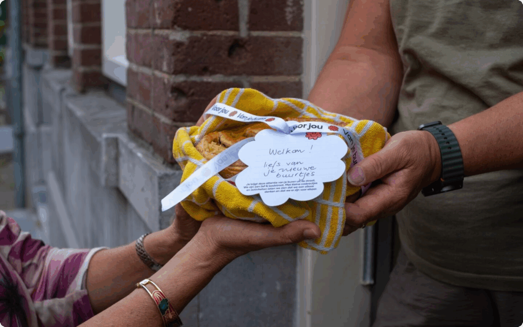 Een buur die een andere buur welkom heet in de straat, je ziet twee paar handen die een buurtcadeau overhandigen.