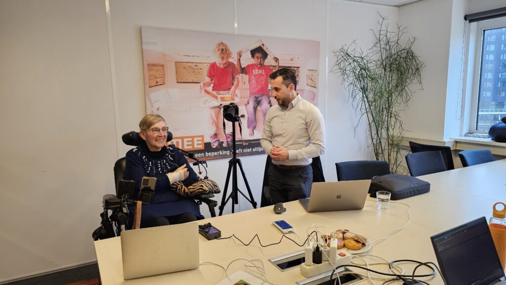 Gebruikerstesten bij CCV payments. Een man in een wit overhemd staat in een vergaderruimte naast een vrouw in een rolstoel. Beiden kijken naar een laptop op de tafel.