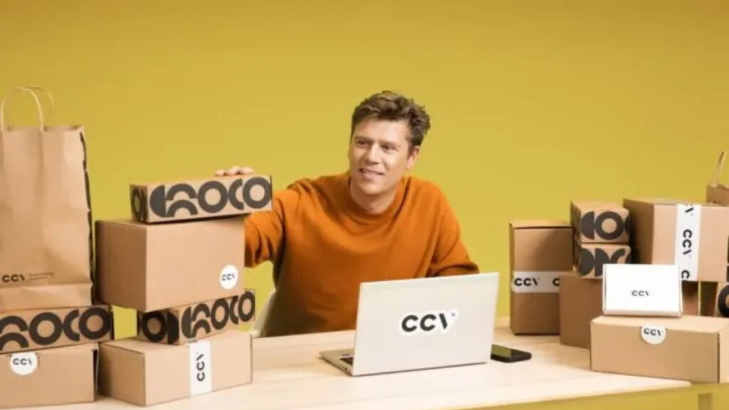 Een ondernemer zit aan een bureau met een laptop met het CCV-logo. Hij is omringd door talrijke kartonnen dozen van verschillende formaten met CCV-branding, tegen een gele achtergrond.