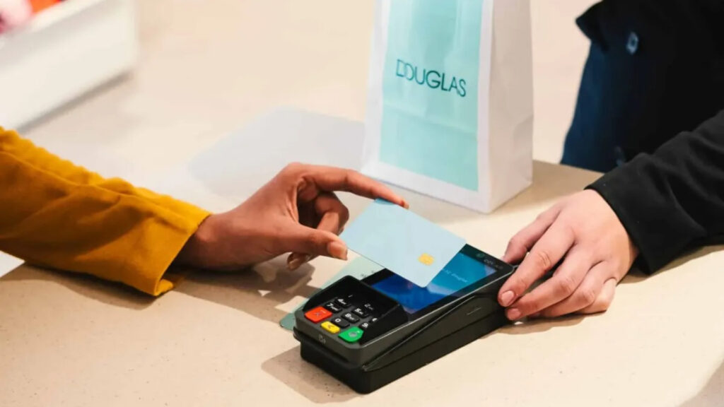 Een close-up van een klant die een lichtblauwe kaart contactloos gebruikt op een zwarte betaalterminal op een toonbank. Op de achtergrond staat een witte papieren draagtas met het Douglas-logo.