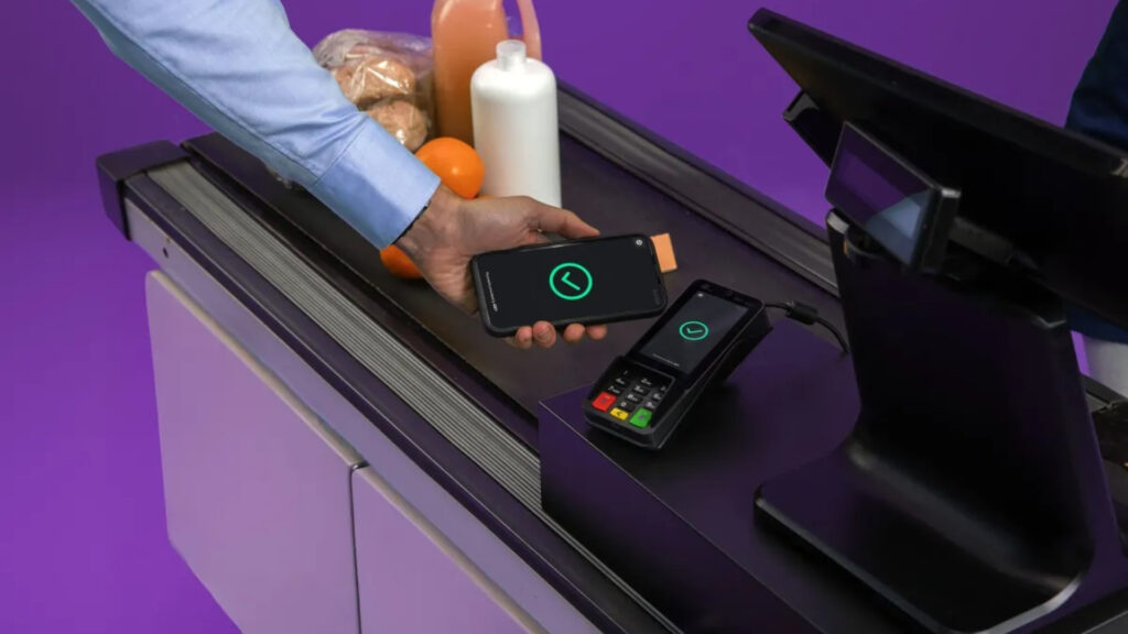 Een klant betaalt bij een supermarkt kassa door een smartphone boven een zwarte betaalterminal te houden. Zowel de telefoon als de terminal tonen een groen vinkje. Op de achtergrond liggen boodschappen zoals sinaasappels en melk tegen een paarse wand.
