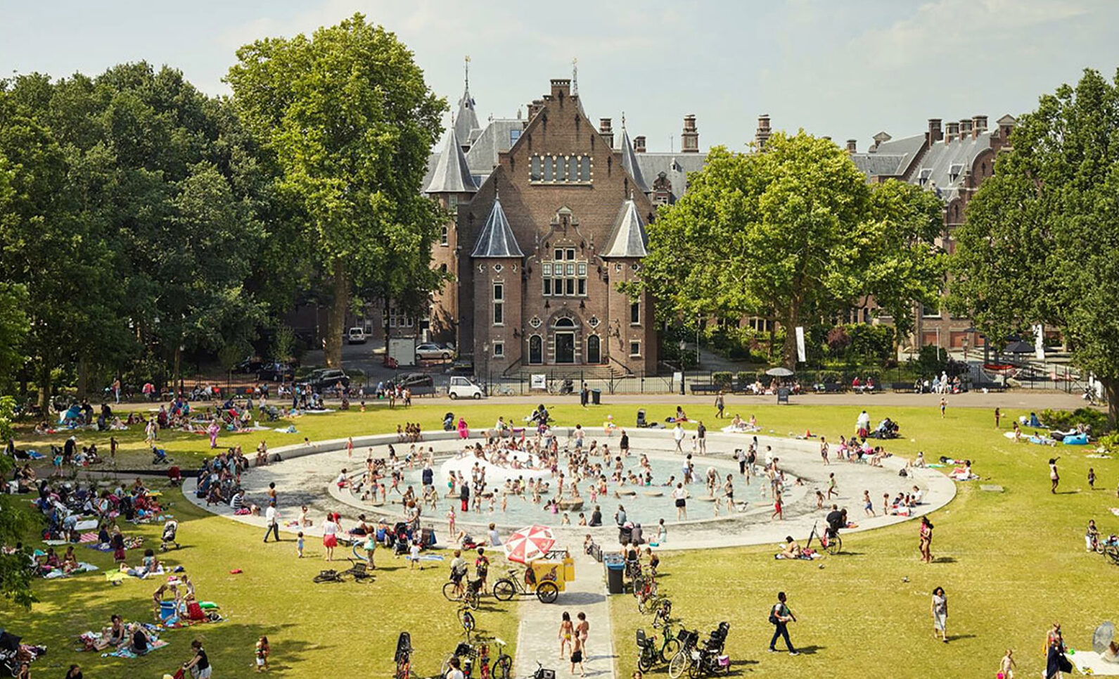 Een bovenaanzicht van de fontein op het grasveld van het Oosterpark, met de voorkant van het Koninklijk Instituut voor de Tropen gebouw op de achtergrond.