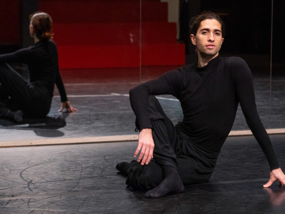 Een danser in zwarte kleding zit op een vloer in een studio met spiegelwand en rode trap op de achtergrond.