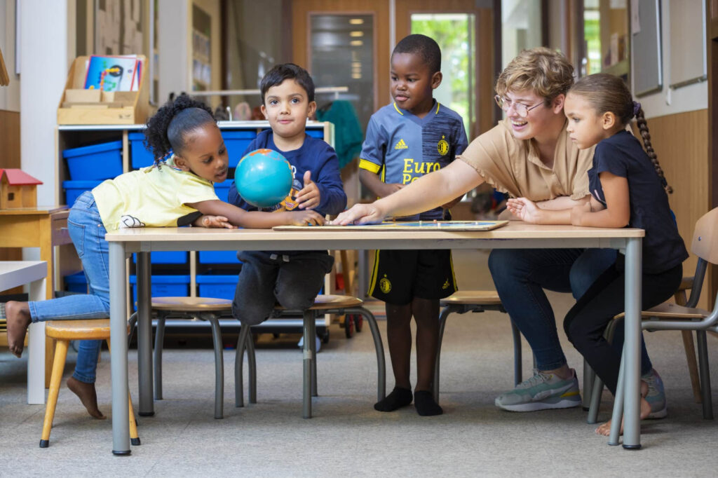 Een leerkracht en vier jonge leerlingen spelen samen aan een tafel in een klaslokaal.