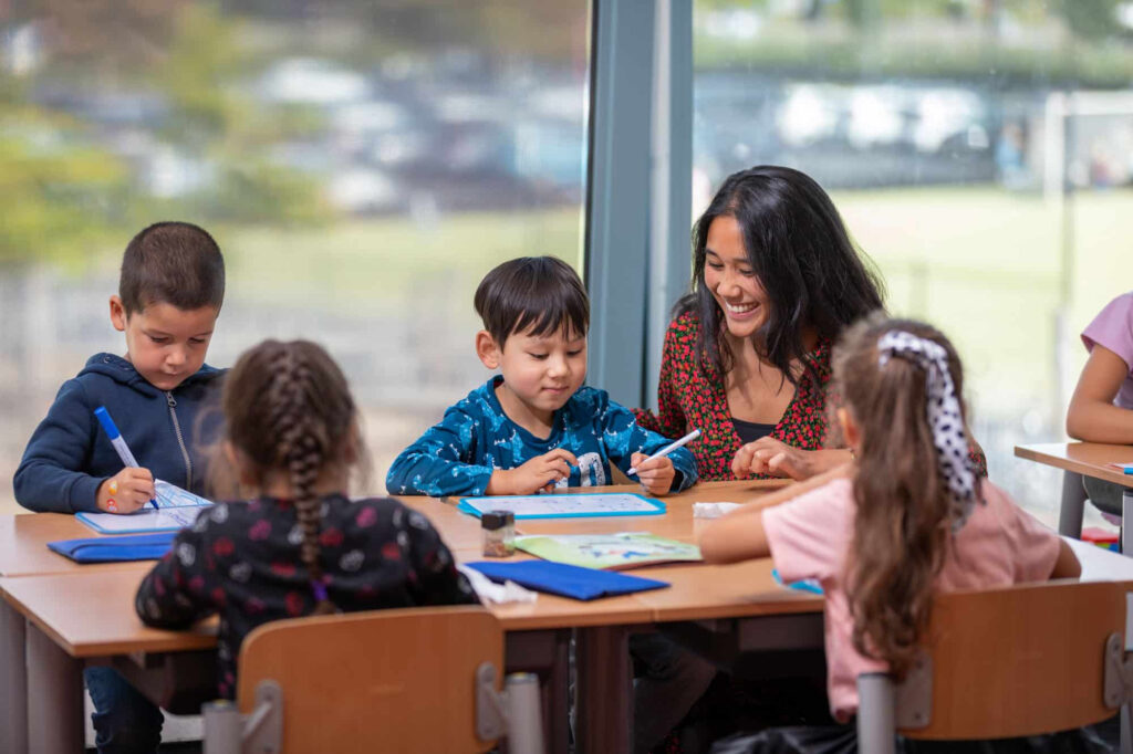 Een lerares glimlacht en helpt vier jonge leerlingen aan een tafel in een klas.