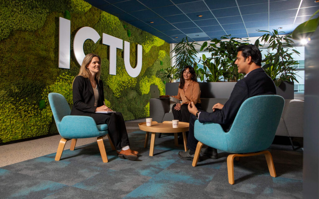 Drie personen in een informele vergaderhoek met een groen mosmuur en het grote ICTU-logo.