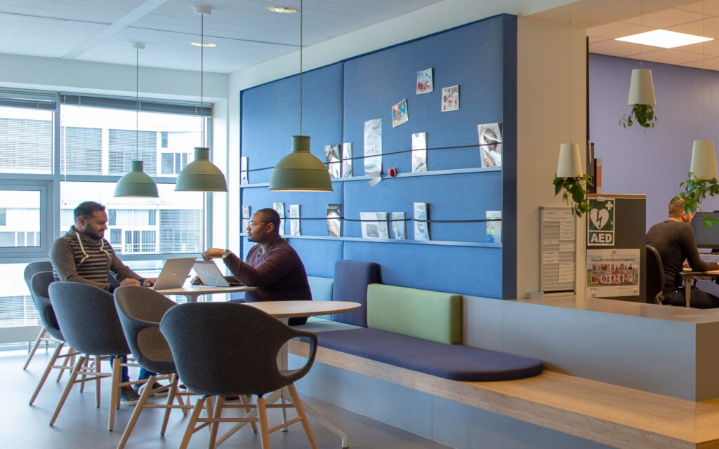 Sfeer foto. Twee mannen aan een tafel met laptops in een informele kantoorzithoek, naast een blauwe prikwand.