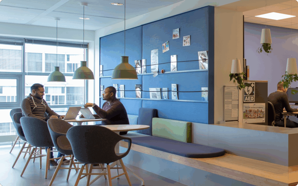 Sfeer foto. Twee mannen aan een tafel met laptops in een informele kantoorzithoek, naast een blauwe prikwand.