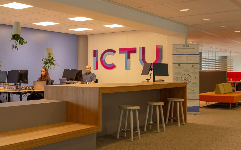 Sfeer foto. Twee medewerkers aan een hoge desk in het ICTU-kantoor met het grote, kleurrijke ICTU-logo op de muur.