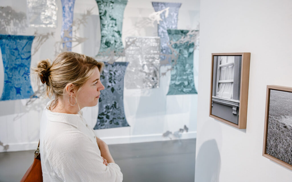 Foto. Vrouw bekijkt kunstwerken in het museum, met een wand vol textiele, sculpturale kunstwerken op de achtergrond.