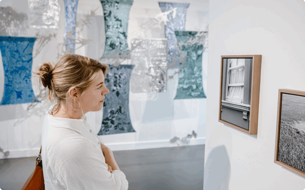 Foto. Vrouw bekijkt kunstwerken in het museum, met een wand vol textiele, sculpturale kunstwerken op de achtergrond.