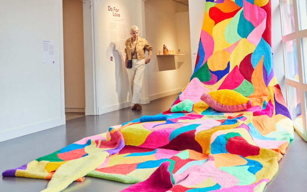 Een felgekleurd, sculpturaal textiel kunstwerk ligt op de vloer van een museumzaal, met een bezoeker op de achtergrond die ernaar kijkt
