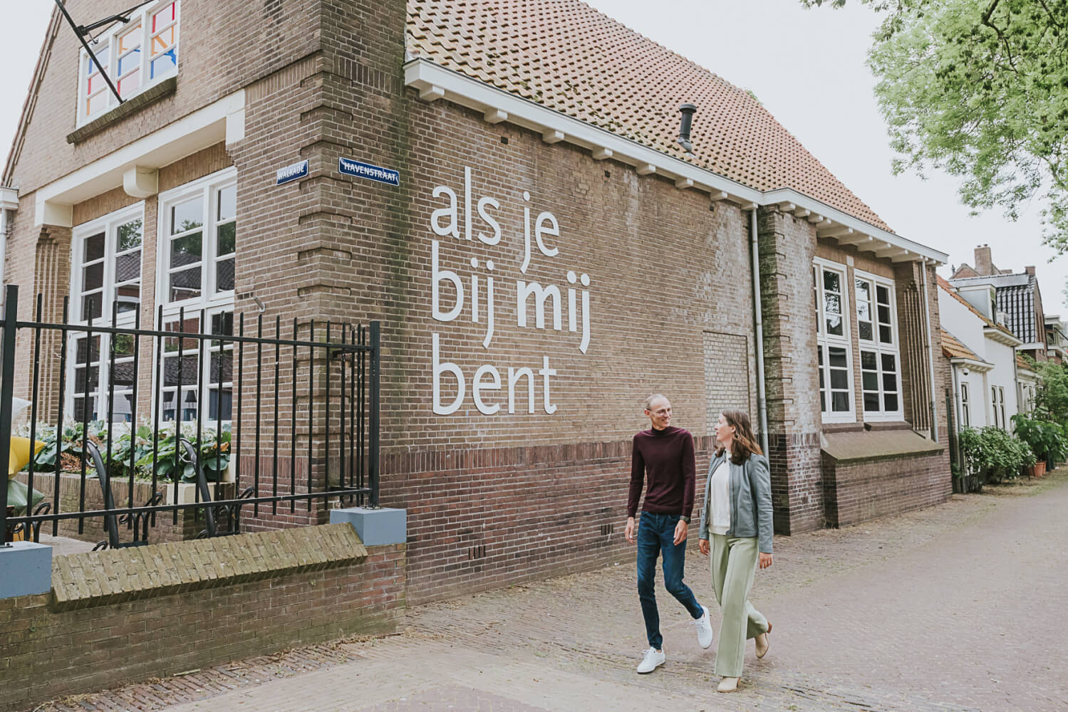 Twee mensen lopen langs het Museum IJsselstein, waar de witte tekst 'als je bij mij bent' op de bakstenen muur staat.