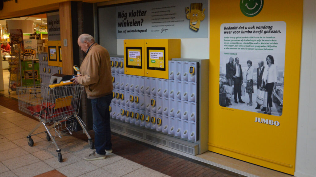 Een oudere man met een winkelwagentje staat bij een zelfscan-station in een Jumbo supermarkt. De wand is gevuld met rijen handscanners en gele informatieborden met de tekst 'Nóg vlotter winkelen?'. Rechts hangt een groot geel bord met een bedankje van de familie van Eerd.