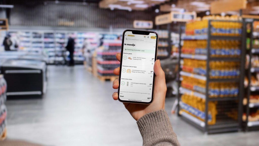 Een persoon houdt een smartphone vast met de Jumbo-app geopend in een wazige supermarktomgeving. Het scherm toont 'Je mandje' met een overzicht van producten zoals brood en kaas, inclusief prijzen en hoeveelheden.