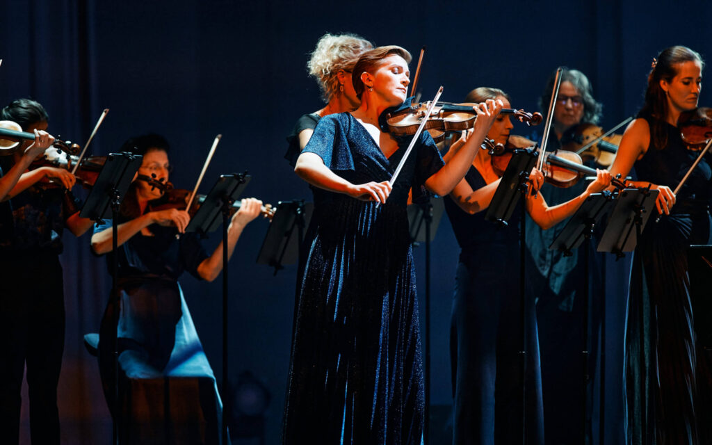 Vrouwelijke musici van het orkest spelen viool in donkerblauwe jurken op een podium met zachte belichting.