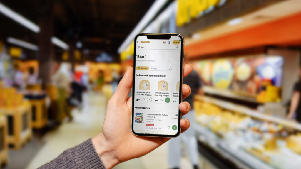 Een close-up van een hand die een smartphone vasthoudt in de kaasafdeling van een Jumbo. Op het scherm is de zoekterm 'Kaas' ingevoerd, met resultaten voor biologische kaas en mozzarella perlines, inclusief afbeeldingen en prijzen.