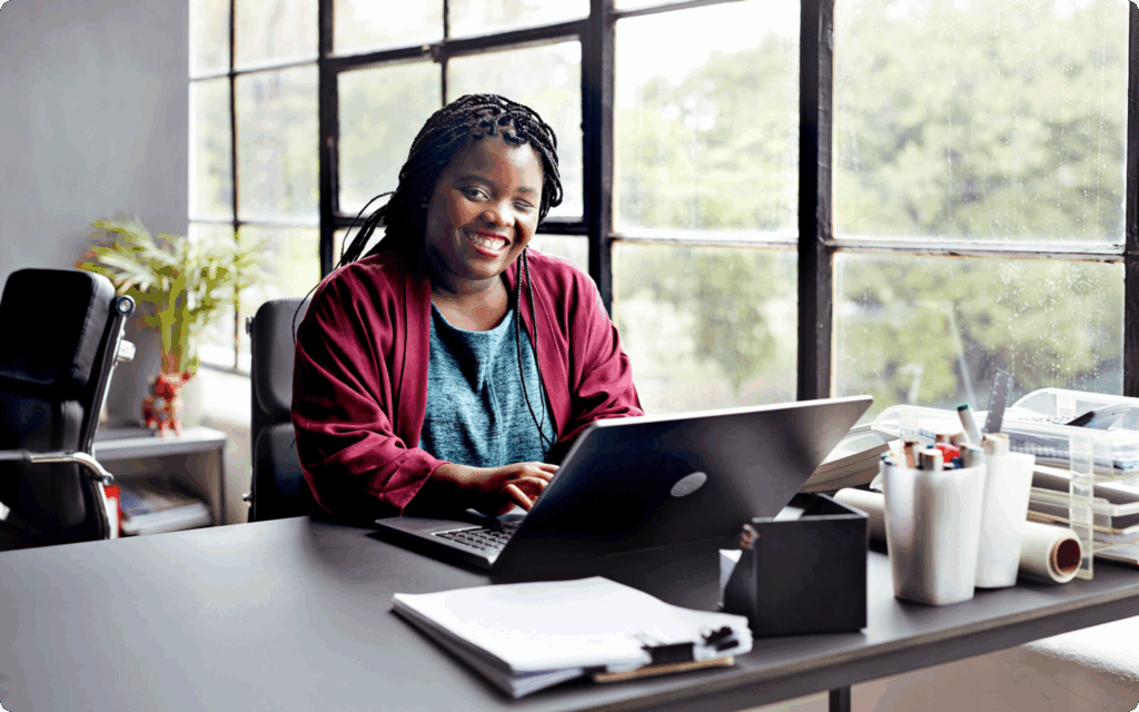Glimlachende vrouw met gevlochten haar zit in een modern kantoor achter haar laptop, met een groot raam.