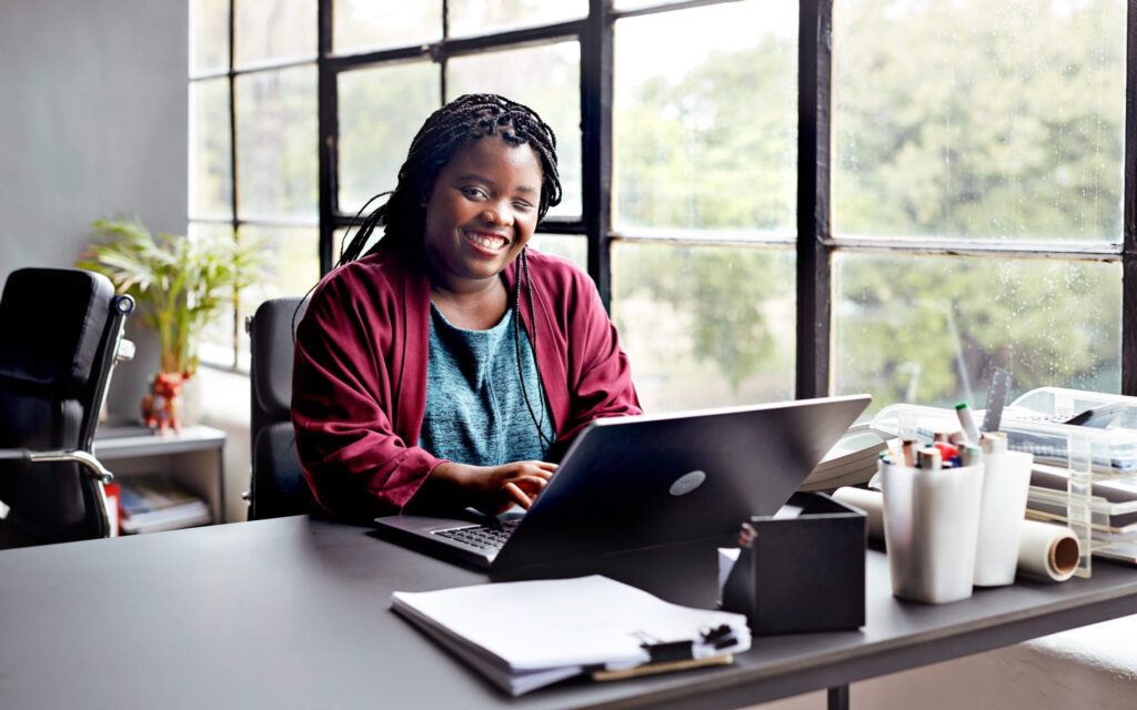 Glimlachende vrouw met gevlochten haar zit in een modern kantoor achter haar laptop, met een groot raam.