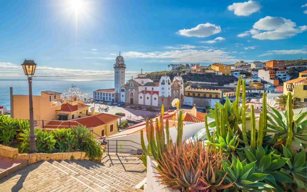 Panoramisch uitzicht op een zonnig mediterraan/Canarisch kuststadje met een witte kerk en helderblauwe lucht.