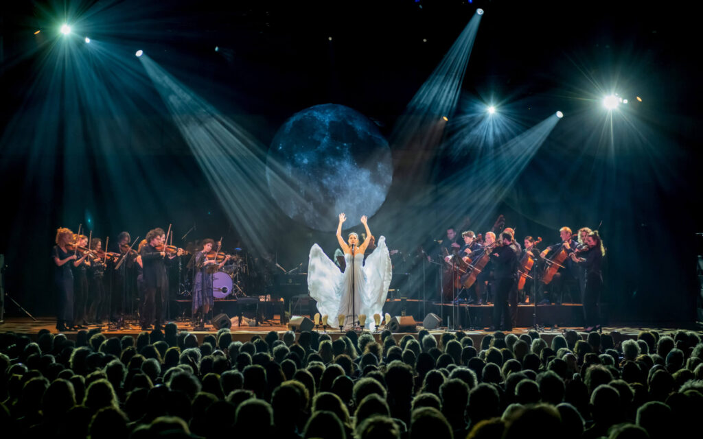 Volledig orkest en een zangeres op het podium met een groot maan-projectie, gezien vanuit een volle zaal.
