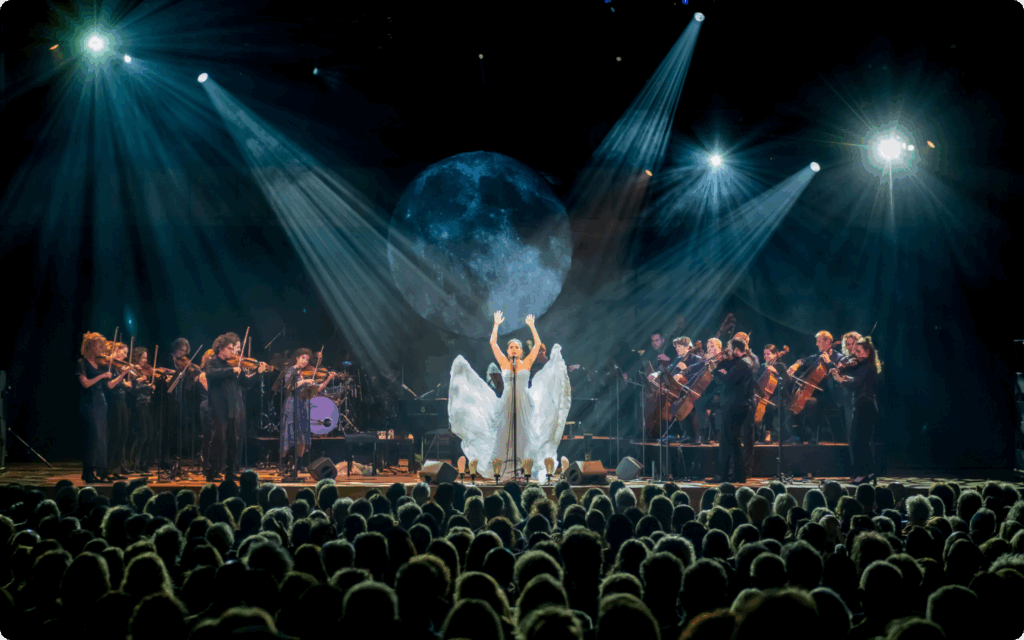 Volledig orkest en een zangeres op het podium met een groot maan-projectie, gezien vanuit een volle zaal.