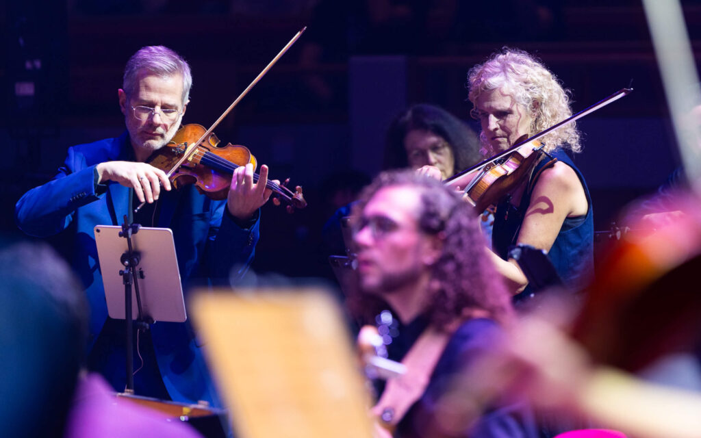 Close-up van musici die viool spelen op het podium, waarbij één muzikant digitale bladmuziek gebruikt op een iPad.