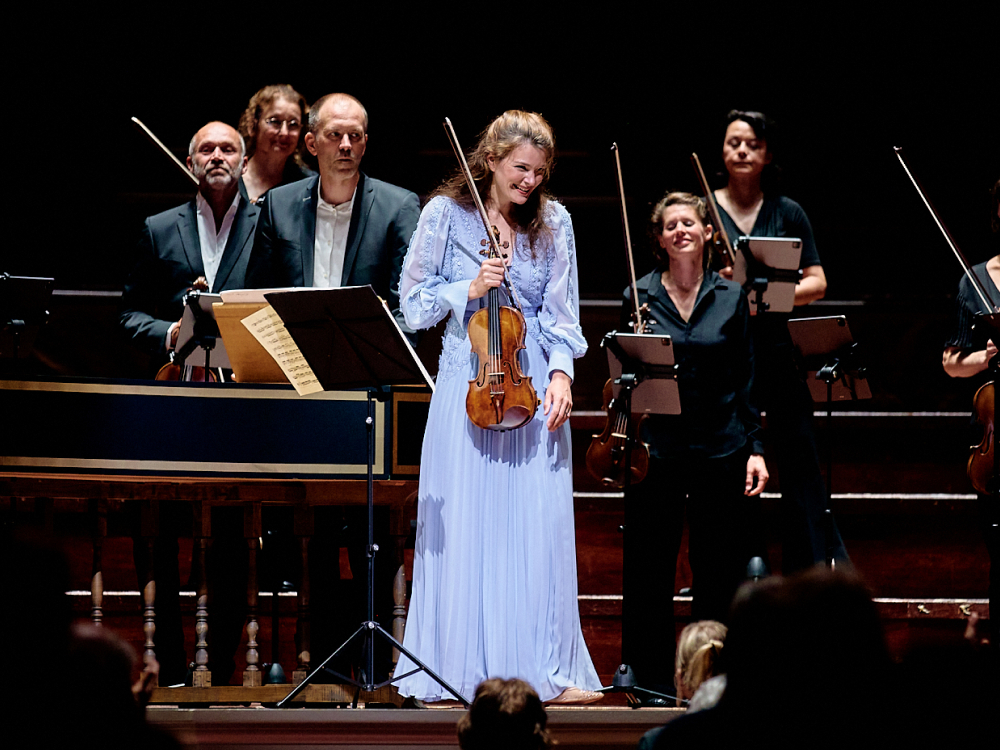 Een vrouwelijke violist met een lange lichtblauwe plooijurk staat glimlachend voor het orkest op het podium.