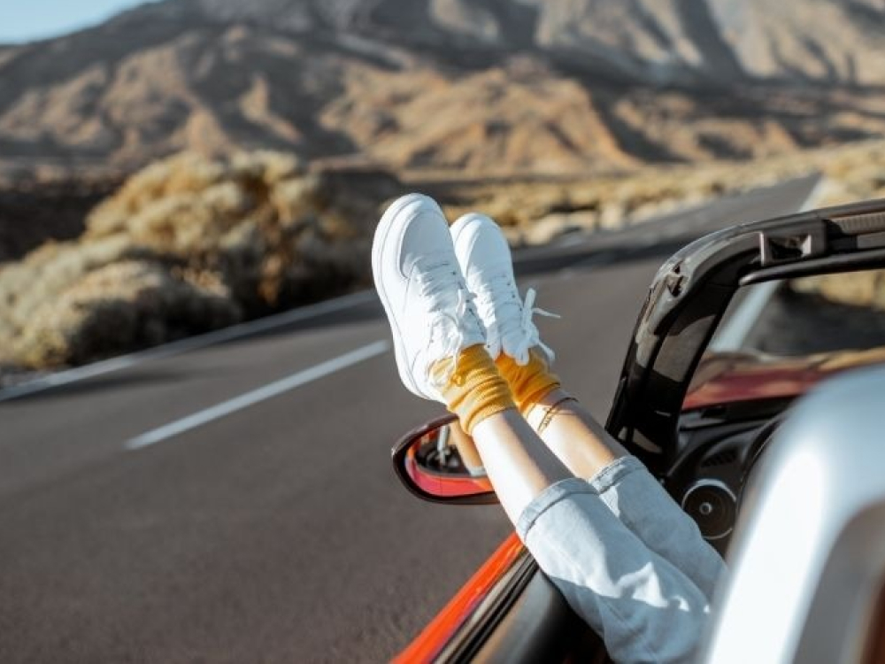 Voeten in witte sneakers steken uit het raam van een auto op een bergachtige weg, wat duidt op een roadtrip.