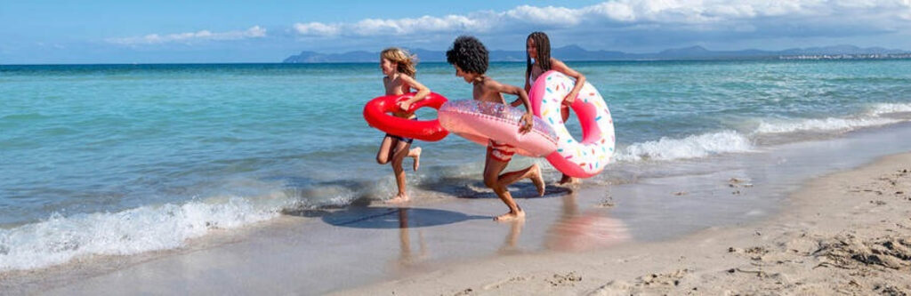 Drie kinderen rennen met zwembanden op een zonnig, breed zandstrand in de branding van de zee.